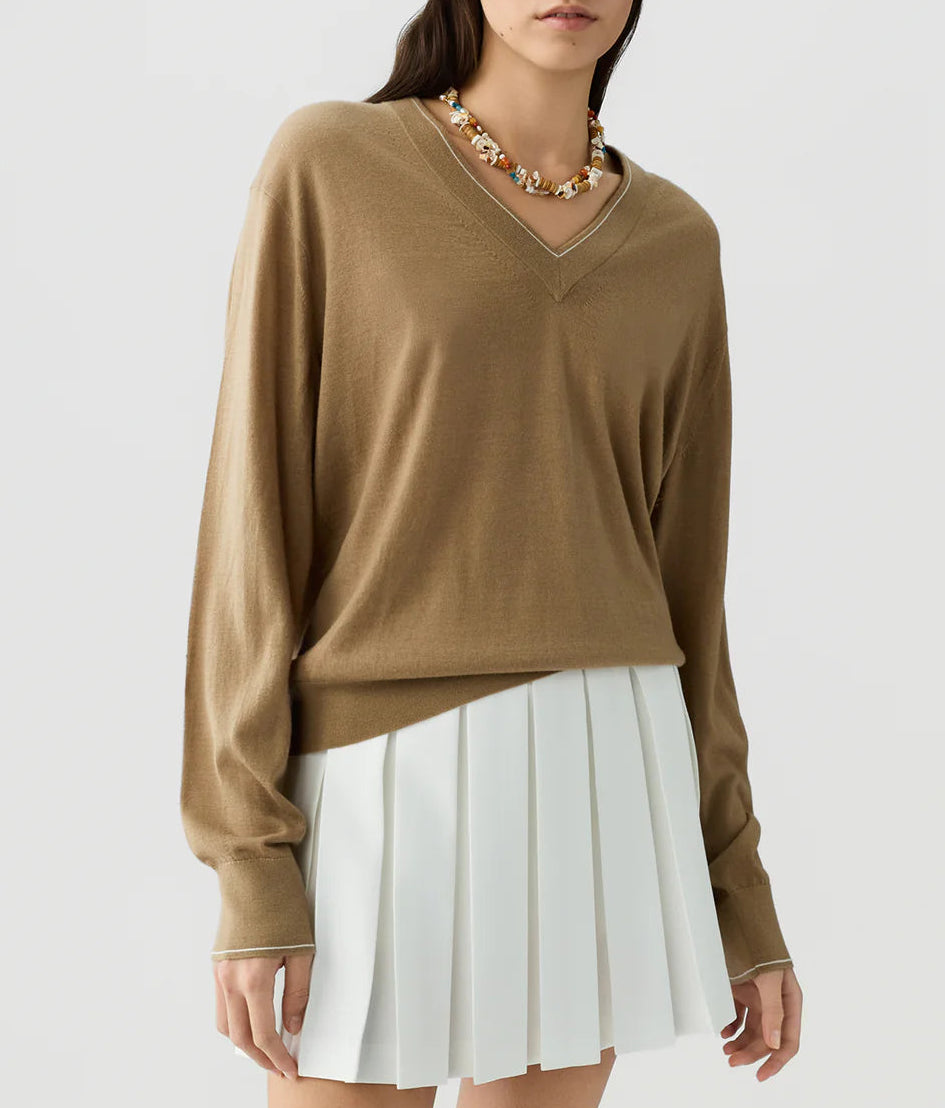 A person wearing The Airy V lightweight cashmere V-neck sweater by Guest in Residence, paired with a white pleated skirt and a beaded necklace, standing against a plain background.