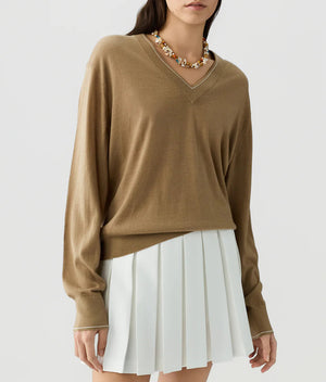 A person wearing The Airy V lightweight cashmere V-neck sweater by Guest in Residence, paired with a white pleated skirt and a beaded necklace, standing against a plain background.