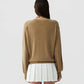A person with long dark hair facing away, wearing the lightweight cashmere "The Airy V" sweater by Guest in Residence in tan and a white pleated skirt, stands against a plain light background.