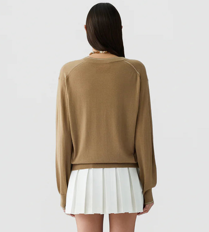 A person with long dark hair facing away, wearing the lightweight cashmere "The Airy V" sweater by Guest in Residence in tan and a white pleated skirt, stands against a plain light background.