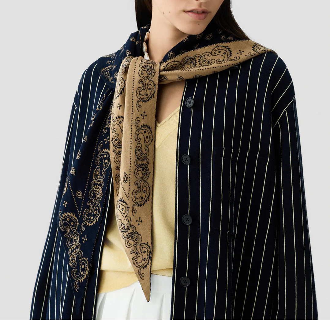 A woman wears a navy pinstripe shirt over a beige top, with The Wild Rag by Guest in Residence—a tan and navy reversible paisley jacquard scarf—draped over her shoulders and knotted at the front.