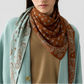 A woman wears a tan top, light blue cardigan, and The Wild Rag—a best-selling two-tone cotton-silk bandana with brown and light blue paisley patterns—from Guest in Residence.