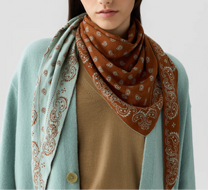 A woman wears a tan top, light blue cardigan, and The Wild Rag—a best-selling two-tone cotton-silk bandana with brown and light blue paisley patterns—from Guest in Residence.