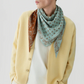 A person wearing a light yellow cardigan, white shirt, and The Wild Rag reversible paisley jacquard scarf in brown and light blue by Guest in Residence, looking to the side against a plain background.