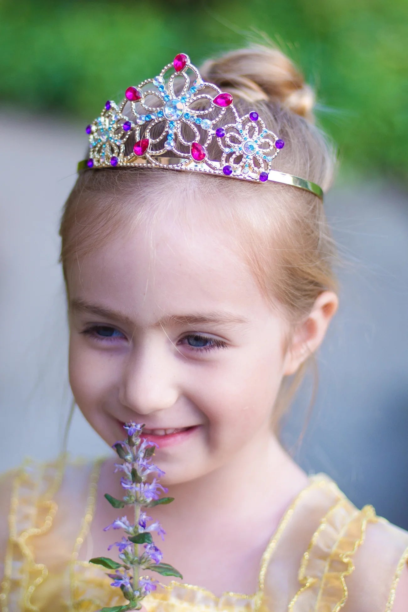 A young girl smiles as she holds a purple flower near her face, wearing the Great Pretenders Bejeweled Tiara Lilac with a shimmering yellow dress and princess costume.