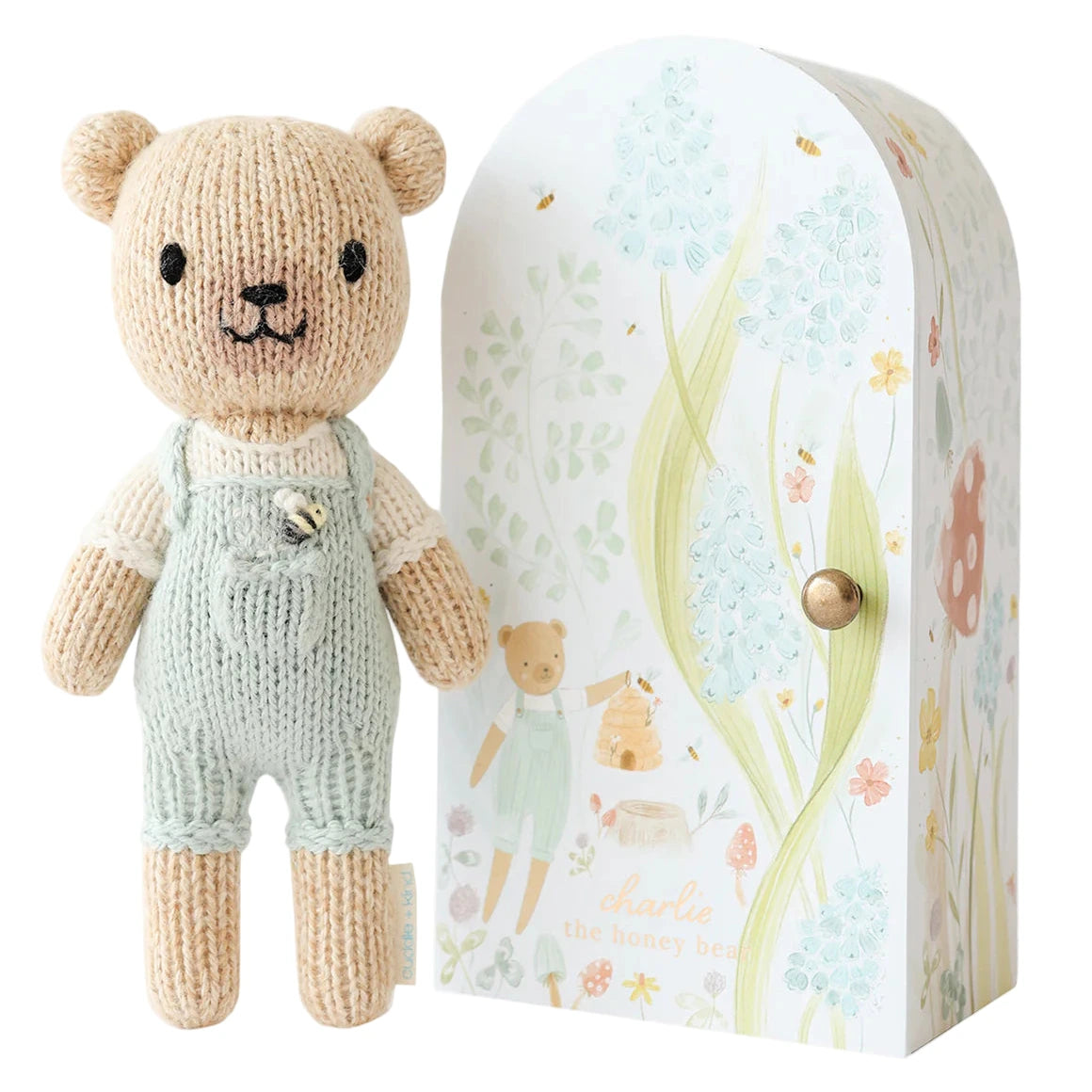 Tiny Charlie the Honey Bear from Cuddle + Kind’s Tiny Collection stands in light green overalls beside a decorative box featuring floral and bear illustrations on a white background.