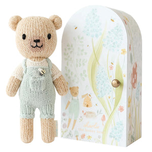 Tiny Charlie the Honey Bear from Cuddle + Kind’s Tiny Collection stands in light green overalls beside a decorative box featuring floral and bear illustrations on a white background.