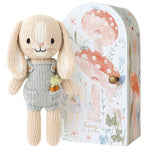 Tiny Henry the Bunny by Cuddle + Kind, a hand-knit doll in gray overalls with a carrot in its pocket, stands beside a decorative box with mushrooms and flowers—part of the fair trade artisan-made Tiny Collection.