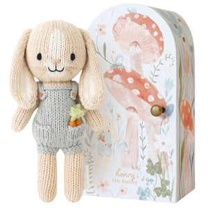 Tiny Henry the Bunny by Cuddle + Kind, a hand-knit doll in gray overalls with a carrot in its pocket, stands beside a decorative box with mushrooms and flowers—part of the fair trade artisan-made Tiny Collection.