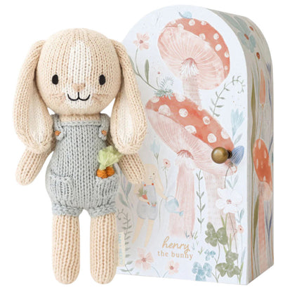 Tiny Henry the Bunny by Cuddle + Kind, a hand-knit doll in gray overalls with a carrot in its pocket, stands beside a decorative box with mushrooms and flowers—part of the fair trade artisan-made Tiny Collection.