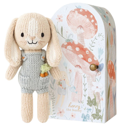 A hand-knit Tiny Hannah the Bunny Blush from Cuddle + Kind, wearing gray overalls, stands beside a decorative box adorned with mushroom and flower illustrations.