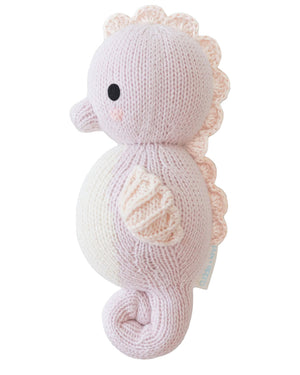 The Baby Seahorse Lilac by Cuddle + Kind is a hand-knit plush in soft pastel hues, made from Peruvian cotton yarn, featuring a round body, curled tail, and scalloped fins—heirloom quality shown side view on white background.