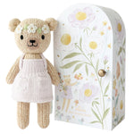 Tiny Olivia the Honey Bear by Cuddle + Kind is a hand-knit bear in a white dress with floral accents, standing beside a matching floral-patterned gift box—an adorable addition to any Cuddle + Kind dolls collection.