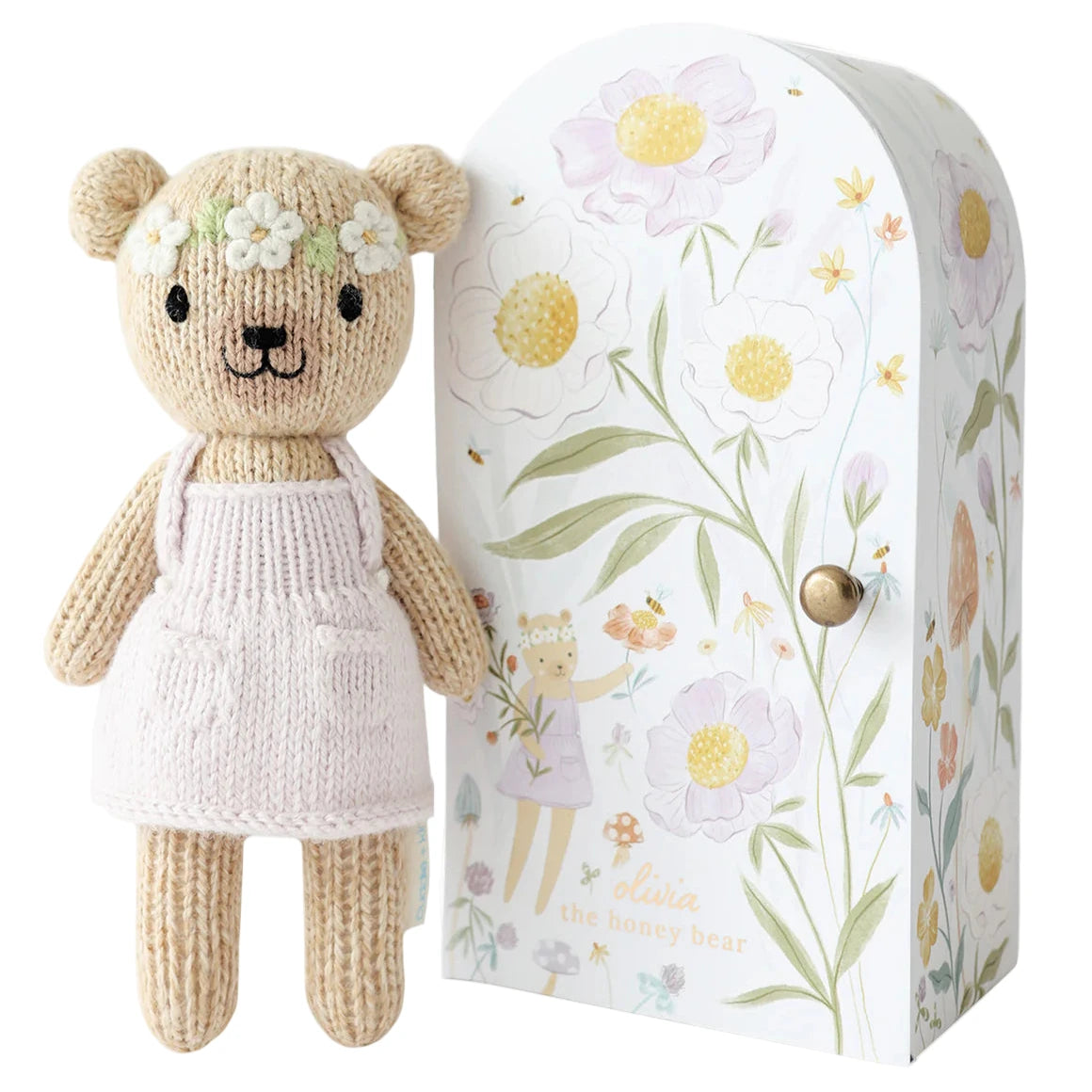 Tiny Olivia the Honey Bear by Cuddle + Kind is a hand-knit bear in a white dress with floral accents, standing beside a matching floral-patterned gift box—an adorable addition to any Cuddle + Kind dolls collection.