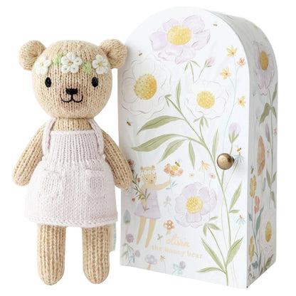 Tiny Olivia the Honey Bear by Cuddle + Kind is a hand-knit bear in a white dress with floral accents, standing beside a matching floral-patterned gift box—an adorable addition to any Cuddle + Kind dolls collection.