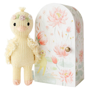 The Tiny Flora the Duckling Mauve by Cuddle + Kind, a knitted yellow plush with floral details, stands beside a floral-patterned box labeled "flora the duckling.