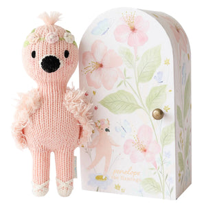 Tiny Penelope the Flamingo by Cuddle + Kind is a hand-knit pink flamingo plush with a flower crown, standing beside a decorative box with floral and flamingo designs, inspired by the brand’s whimsical fair trade dolls.