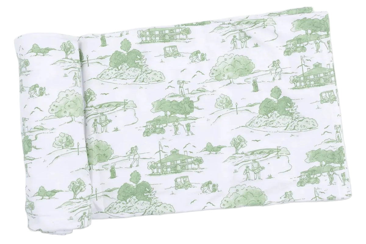 White fabric adorned with a green pastoral toile design of houses, trees, and people outdoors, partially unrolled on a white background—this Angel Dear Swaddle Blanket combines charming style with cozy comfort for your baby.
