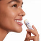 A woman smiles, holding Supergoop Round Trip Ready near her lips—perfect for adding a lightweight, travel-friendly sunscreen to your SPF kit.