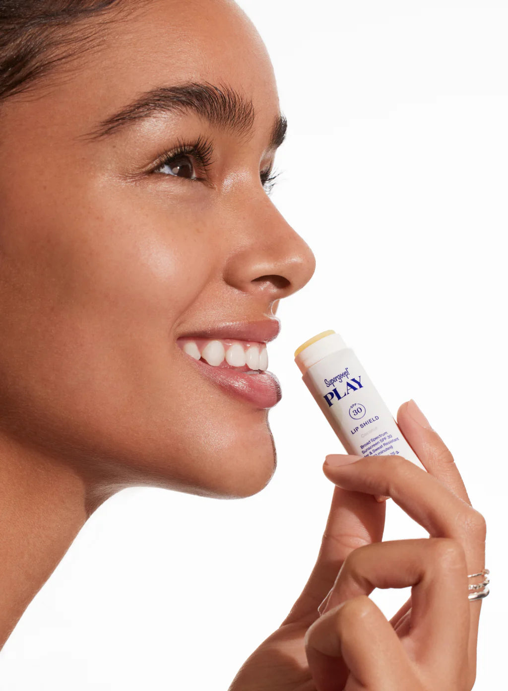 A woman smiles, holding Supergoop Round Trip Ready near her lips—perfect for adding a lightweight, travel-friendly sunscreen to your SPF kit.