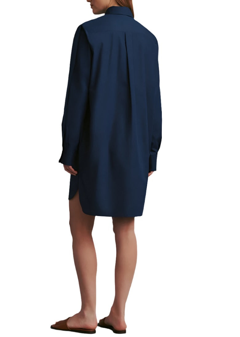 A person wearing a dark blue, long-sleeved TWP Ma House Dress and brown flat shoes, standing and facing away. The outfit subtly blends the casual comfort of an oversized shirt with the elegance of a summer dress by TWP.