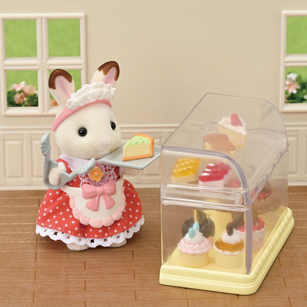 A Calico Critters toy rabbit in a red dress holds a slice of cake with tongs beside a display case brimming with beautifully detailed sponge cakes and assorted toy desserts, all part of the Village Cake Shop Starter Set—perfect for imaginative play and decorating your own cake shop scenes in a cozy room with two windows.