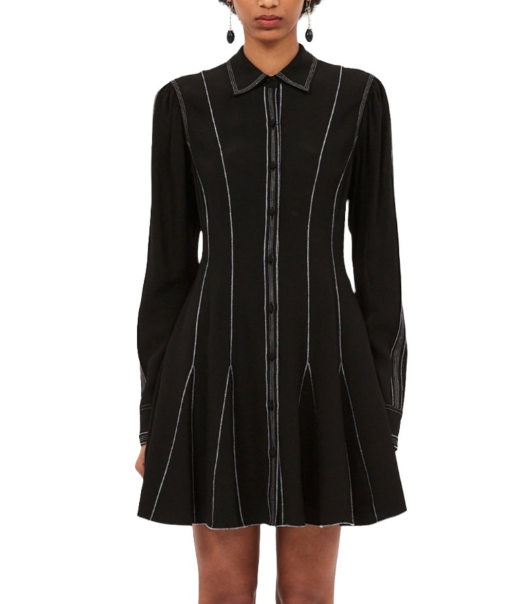 A person wearing an Ulla Johnson Roanne Dress, which features a black long-sleeve shirt-inspired mini silhouette with white vertical stitching and a collar, stands facing forward.