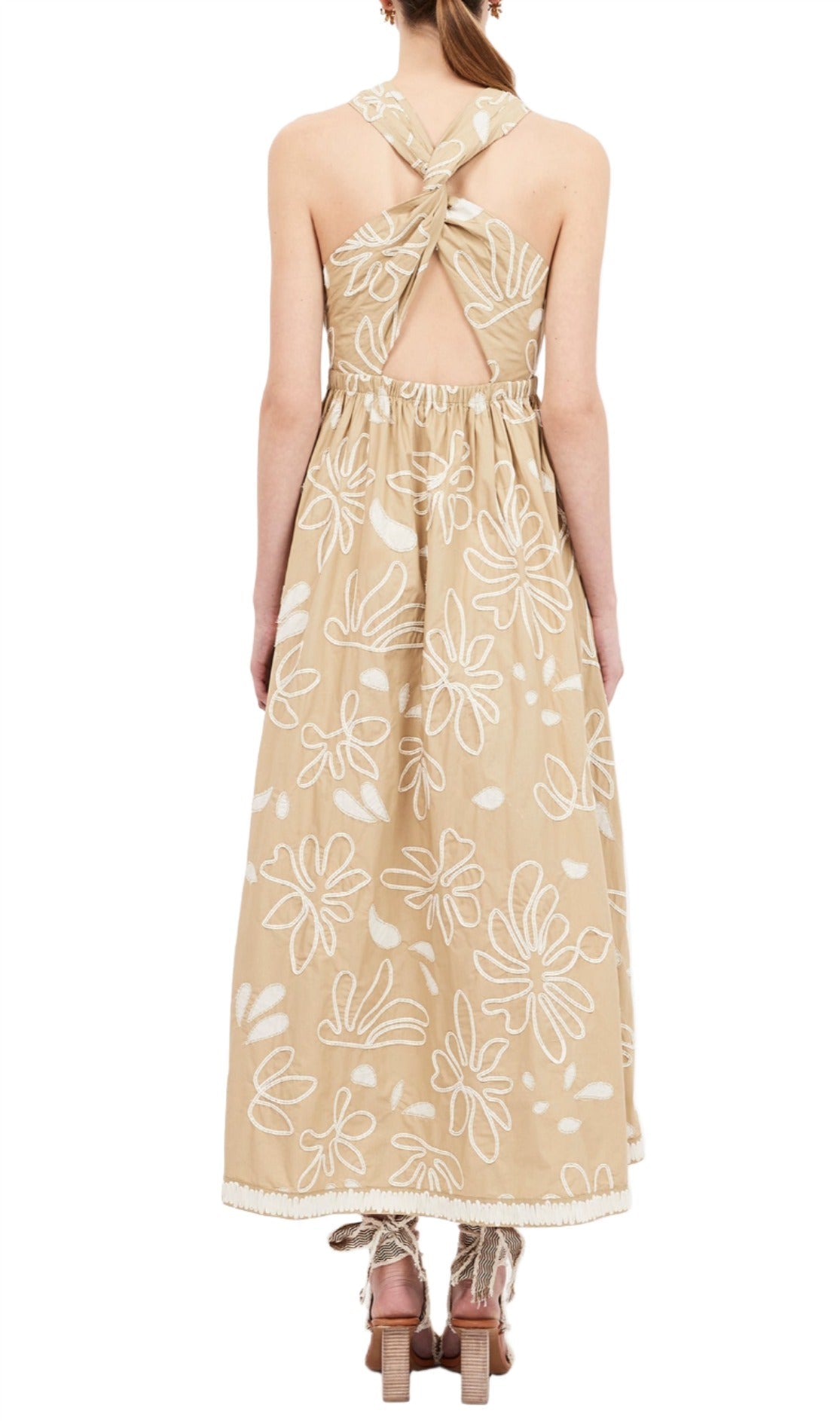 Back view of a woman wearing the Ulla Johnson Viola Dress, a tan sleeveless dress with embroidered florals, crisscross straps, and a gathered waist, standing on beige platform heels.