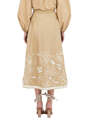 Rear view of a person wearing the Ulla Johnson Layla Skirt, a beige A-line skirt adorned with white floral embroidery, paired with a coordinating top featuring puffed sleeves, and strappy high-heeled sandals.