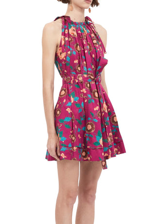 A person wearing a sleeveless, pink floral-patterned Ulla Johnson Brienne Dress with a halter neck and a cinched waist. The poplin dress features pleated details and a mini length skirt. The person's face is not visible.