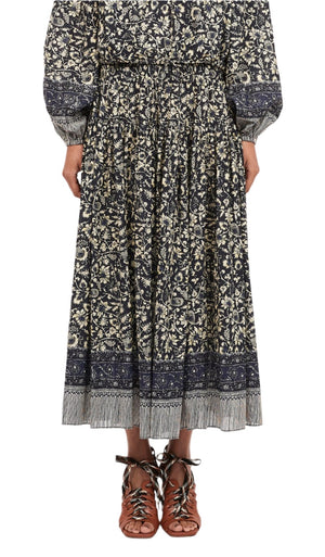Standing against a white background, the person wears an Ulla Johnson Avia Skirt featuring a long, patterned black and white floral design reminiscent of a vintage batik print. Paired with brown lace-up sandals, the ensemble captures timeless elegance.