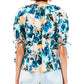A woman elegantly models the Ulla Johnson Vienne Blouse, showcasing its vibrant brushstroke pattern and 100% cotton fabric with elbow-length tie sleeves, paired with light-colored jeans, from a back view.