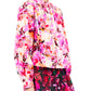 The individual is styled in Ulla Johnson's Tahlia Blouse featuring a floral print, long sleeves, and collar, paired with a matching cotton skirt against a white backdrop. The look is accentuated by artistic brushstroke pink earrings.