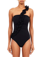 A person wearing the Ulla Johnson Capri Maillot One Piece, featuring a black one-shoulder design with an elegant asymmetric ruffle strap cascading across the front.