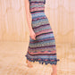 A woman in the Ulla Johnson Ajak Knit Dress—a sleeveless, multicolored striped, rib-knitted midi with ruffled edges—stands on a patterned floor against a textured beige backdrop.