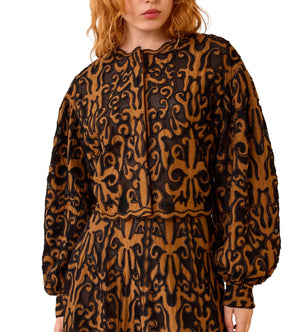 A woman wears the Ulla Johnson Coletta Applique Jacket, featuring a long-sleeved, brown and black pattern with intricate, ornate details reminiscent of Victorian embroidery.