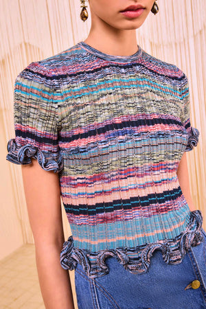 A woman wears the Ulla Johnson Miya Knit Top, a short-sleeve piece made from multicolored space-dyed yarns with striped and ruffled sleeve and hem details, paired with high-waisted blue jeans.
