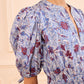 A woman stands against a cream-colored background wearing the Ulla Johnson Adriana Dress—crafted from cotton poplin, with blue and red abstract designs, puffed sleeves, and a gathered waist.