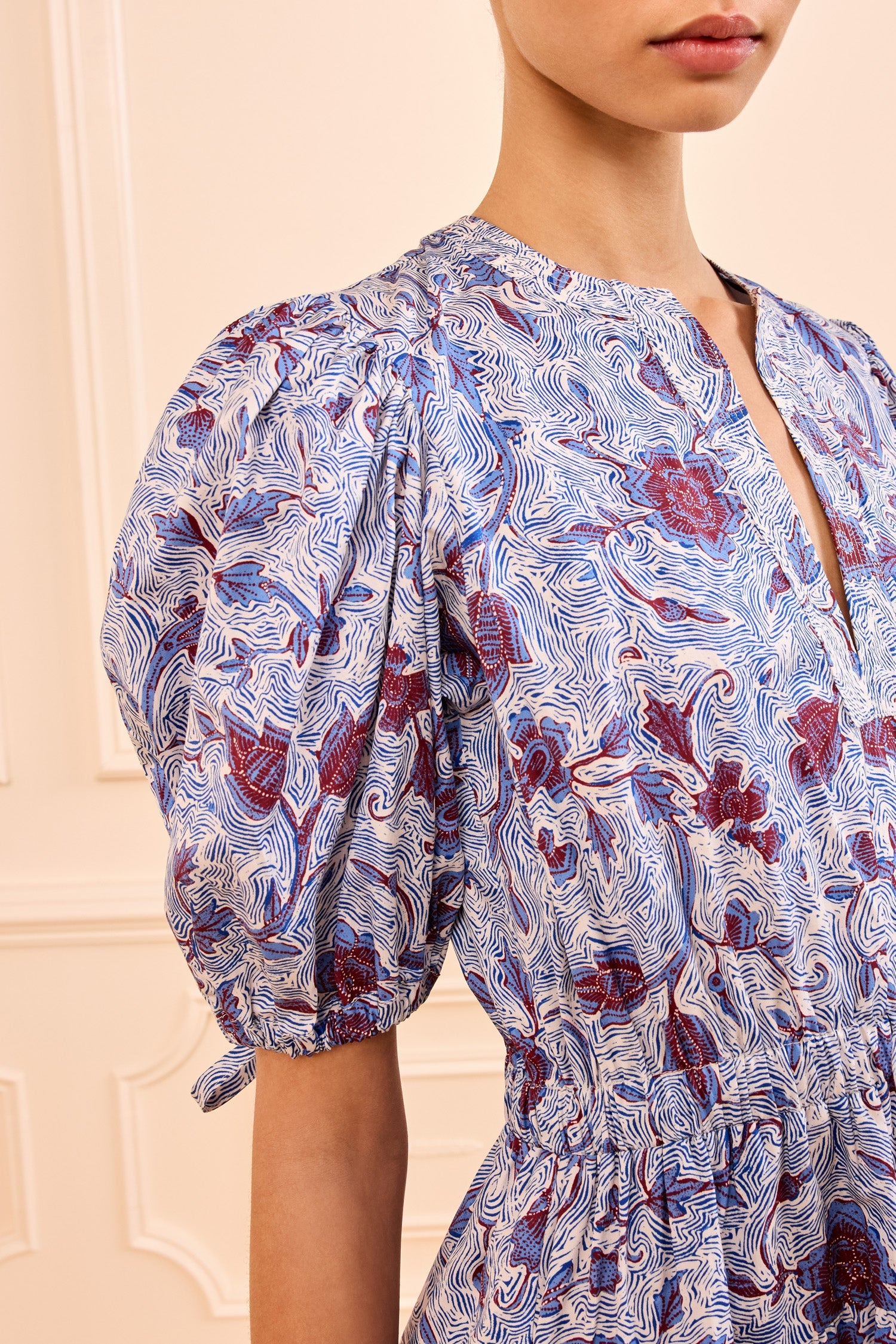 A woman stands against a cream-colored background wearing the Ulla Johnson Adriana Dress—crafted from cotton poplin, with blue and red abstract designs, puffed sleeves, and a gathered waist.