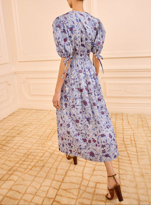 A person wearing the Ulla Johnson Adriana Dress in blue and white cotton poplin with puff sleeves and brown heeled sandals stands in a beige carpeted room.