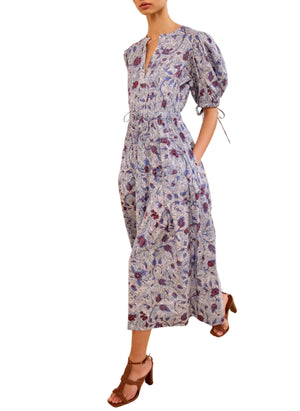 A person wearing the Ulla Johnson Adriana Dress—an ankle-length cotton poplin dress with a light blue floral print, puff sleeves, V-neck, cinched waist—and brown heeled sandals.