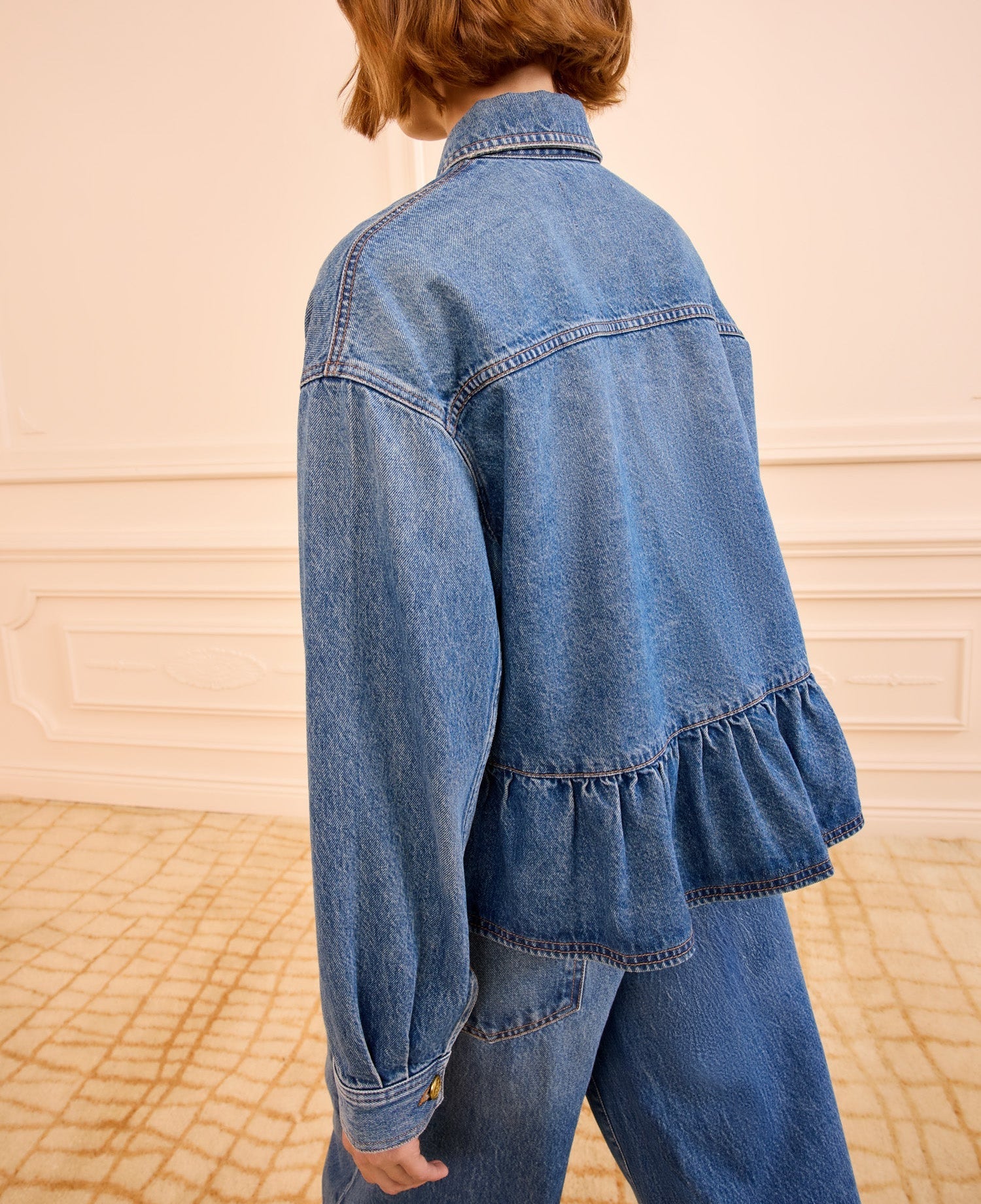 Wearing Ulla Johnson's blue denim Arquette Jacket with a ruffled hem and matching pants, this person stands indoors on a patterned carpet.