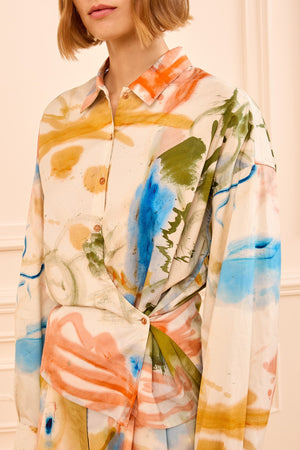 A woman wears the Ulla Johnson Artist Blouse, a collared long-sleeve top in satin-faced poplin with an abstract multicolored print featuring orange, blue, green, and beige tones.