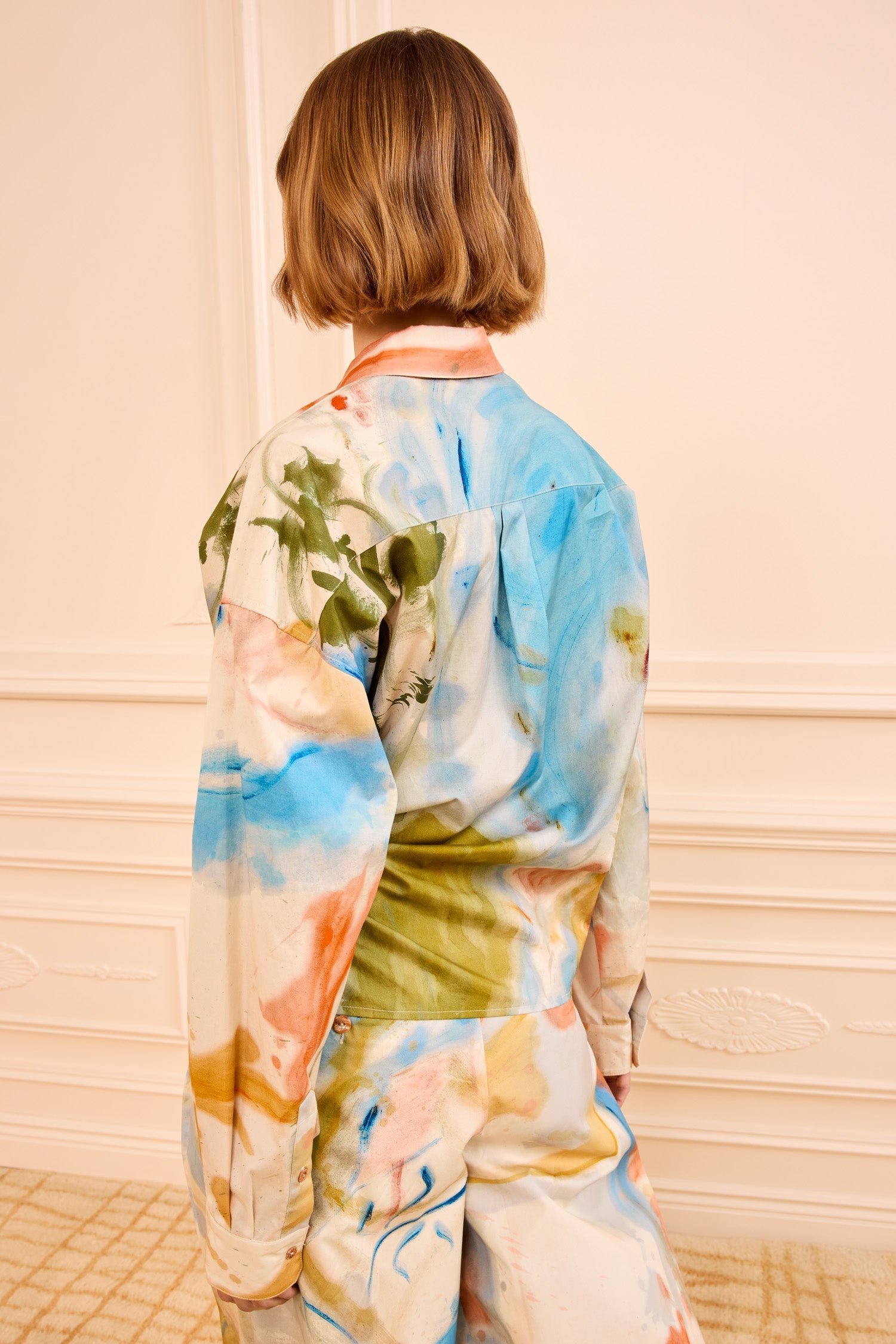 A person with short, light brown hair faces away, wearing the Ulla Johnson Artist Blouse— a loose, long-sleeved top in satin-faced poplin featuring a colorful abstract pattern.