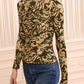 A person wearing the Ulla Johnson Asha Jersey Top—a fitted, long-sleeve crewneck with green and gold floral print and gathered detailing—paired with blue jeans stands in front of a cream paneled wall.