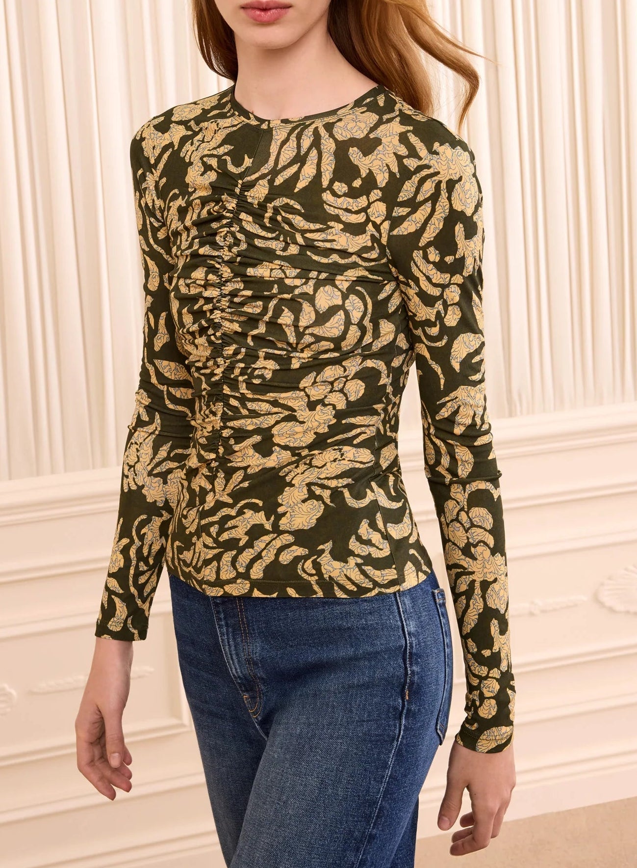 A person wearing the Ulla Johnson Asha Jersey Top—a fitted, long-sleeve crewneck with green and gold floral print and gathered detailing—paired with blue jeans stands in front of a cream paneled wall.