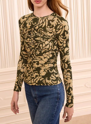 A person wearing the Ulla Johnson Asha Jersey Top—a fitted, long-sleeve crewneck with green and gold floral print and gathered detailing—paired with blue jeans stands in front of a cream paneled wall.