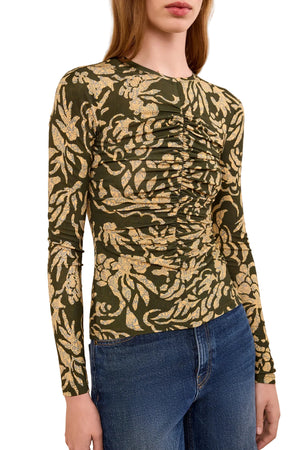 A woman wears the Ulla Johnson Asha Jersey Top, a ruched long-sleeve crewneck featuring a green and yellow leaf pattern, paired with blue jeans.