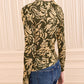 Wearing the Ulla Johnson Asha Jersey Top—a fitted, long-sleeve green jersey adorned with a gold floral pattern and gathered cuffs—paired with blue jeans, a person stands in front of a cream-colored paneled wall.