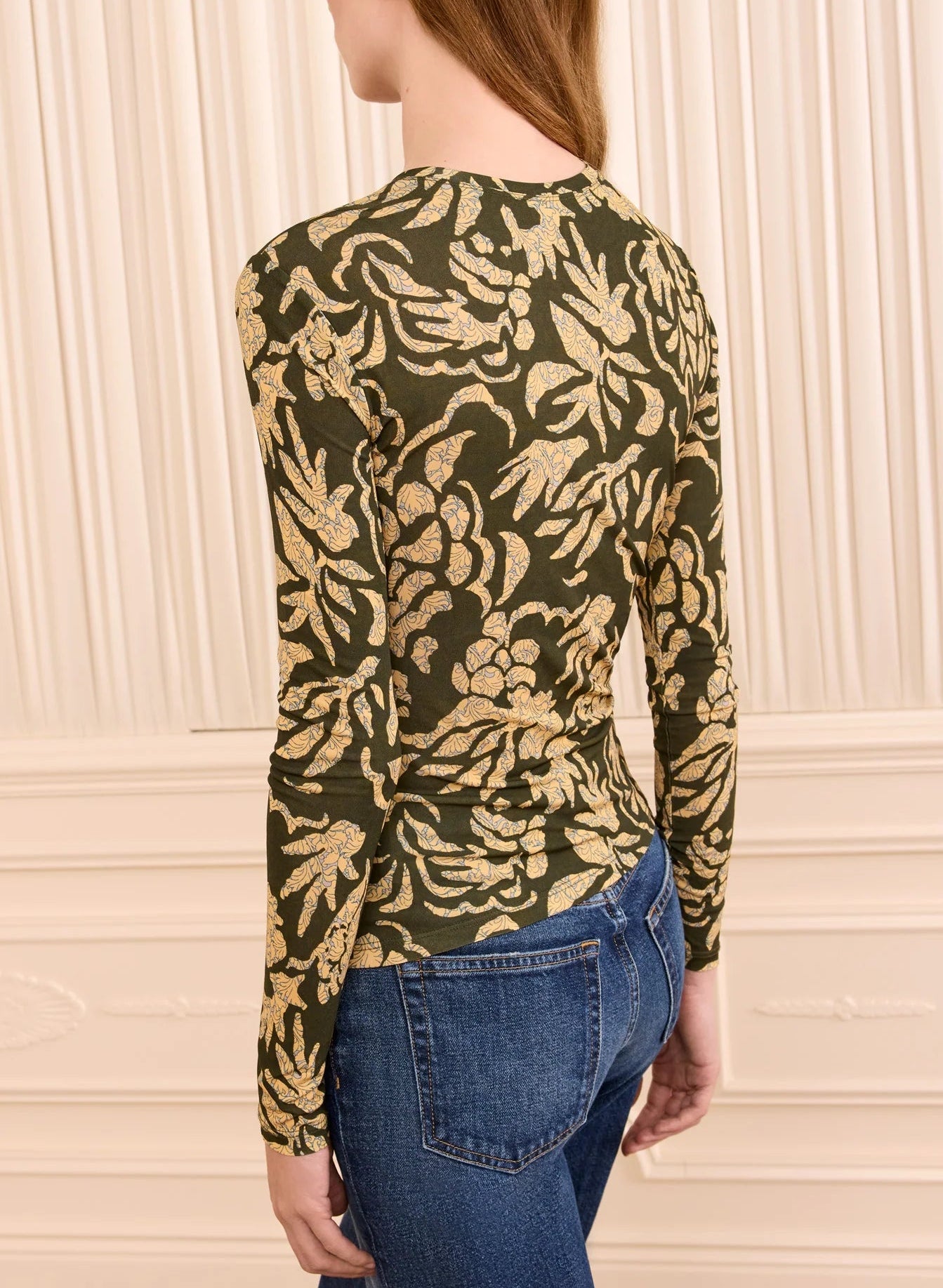 Wearing the Ulla Johnson Asha Jersey Top—a fitted, long-sleeve green jersey adorned with a gold floral pattern and gathered cuffs—paired with blue jeans, a person stands in front of a cream-colored paneled wall.